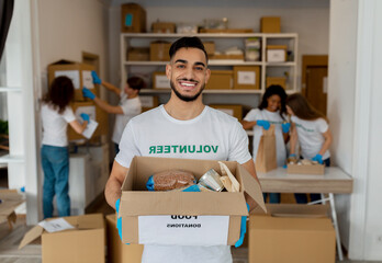A male volunteer at a charity center holds a box filled with food items. He works alongside colleagues, organizing donations to support poor and elderly individuals in the community.