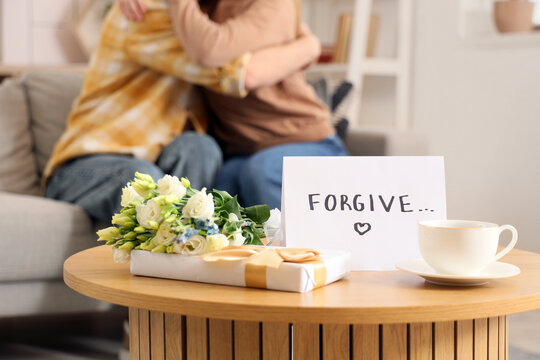 Paper with word FORGIVE, coffee cup and gifts on table against hugging couple at home, closeup