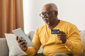 An older man sits comfortably on a couch, actively shopping online with his tablet while holding a credit card. The warm living room setting creates a cozy atmosphere.