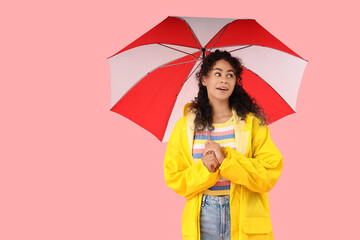 Young African-American woman in raincoat with umbrella on pink background