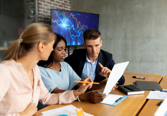 Colleagues from different backgrounds discuss financial reports in a contemporary office. They are seated at a table, reviewing documents and sharing insights during a collaborative meeting.