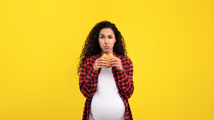 A woman in a plaid shirt stands holding a burger with a thoughtful expression. She is visibly pregnant and poses against a vibrant yellow backdrop, adding a playful touch to the scene. © Prostock-studio