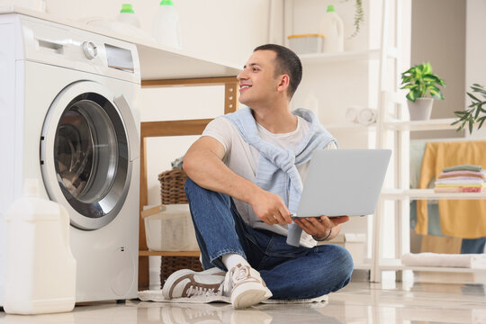 Young man using laptop near washing machine in laundry room