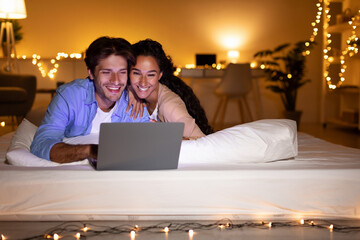 Married couple lies in bed at night, using a laptop to watch a film. They are relaxed and enjoying...