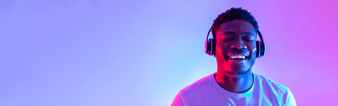 A handsome young black man with headphones smiles joyfully while listening to music. He has his eyes closed, immersed in the rhythm, surrounded by colorful neon lights.