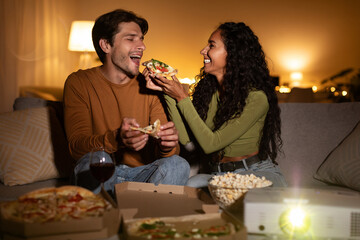 Cheerful couple sits on the couch feeding each other slices of pizza while watching a film at home. The cozy atmosphere is enhanced by the home cinema projector and soft lighting.
