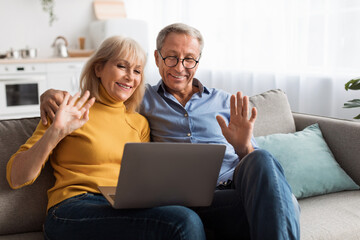 An elderly couple sits closely together on a couch, smiling and waving at their laptop screen during a joyful video call. The bright, cozy living room adds to the warmth of their moment.