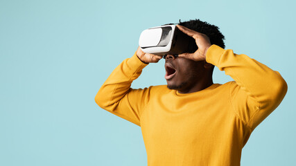 Excited young black man enjoying virtual reality glasses experience, looking towards copy space with mouth open, modern entertainment concept, blue studio background, panorama