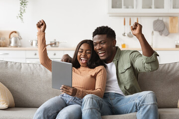 Happy african american couple sits on a couch, playing online games on a tablet. They share joyful moments, celebrating victories as they engage in friendly competition at home.