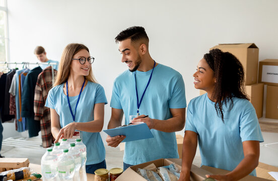 An Arab male manager checks a donation list and writes on a clipboard while diverse volunteers work together to pack donation boxes in a community center, supporting a charitable organization.
