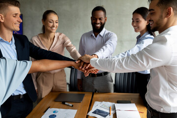 Group of diverse colleagues in a corporate office come together to celebrate their success by stacking hands. This moment showcases unity and collaboration among team members during a meeting.