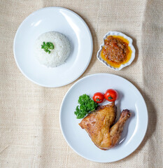Top view of roasted chicken leg served with steamed white rice, chili sauce, and garnish on white plates. Clean flat lay food image with copy space, ideal for Asian cuisine, menu, or lifestyle content