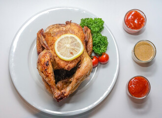Top view of whole roasted chicken served on white plate with lemon slice, parsley, cherry tomatoes, and dipping sauces. Clean commercial food image for menus, ads, and recipes