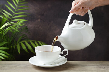 Woman pouring hot tea into cup from teapot at wooden table, closeup. Space for text