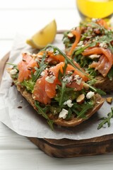 Tasty bruschetta with salmon, avocado, arugula, seeds and feta cheese on white wooden table, closeup