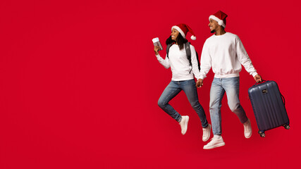 Joyful duo dressed in holiday sweaters and Santa hats hold hands while preparing for a trip. They are carrying luggage and enjoying the festive spirit, all against a vibrant red backdrop.