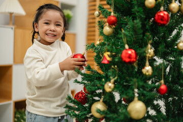 toddler girl decorating toy ball on Christmas tree at home