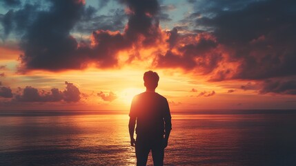 Fototapeta premium A man stands silhouetted against a vibrant sunset over a calm ocean, the sky ablaze with fiery orange and pink hues.