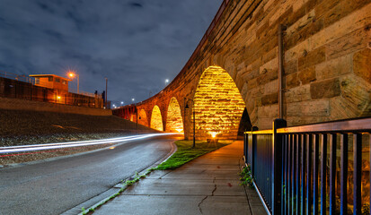Stone Arch Bridge features illuminated arches at night in Minneapolis. Long exposure captures light trails on West River Parkway under a cloudy sky