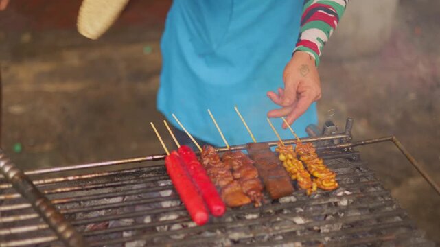 Night market grilling, vendor basting skewers amidst festival atmosphere, chef turning skewers over hot coals at night market, asian vendor in straw hat tending to meat on open flame at festival stall