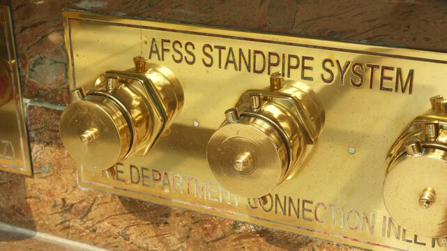 Urban brass standpipe panel, masonrymounted brass panel with capped outlets and water residue, reflective brass fire safety panel displaying capped outlets on urban masonry wall with signs of wear