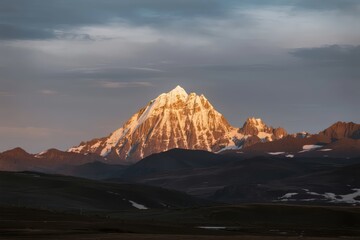 Snow-capped mountain peak illuminated by golden light at dawn, surrounded by rugged terrain and distant ridges under a moody sky