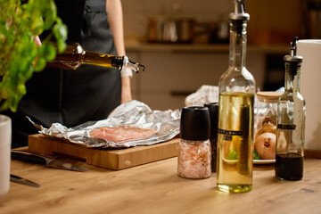 Close-up of woman pouring olive oil on raw chicken breast with seasoning on foil in home kitchen. Concept of food preparation, cooking process and homemade meal.