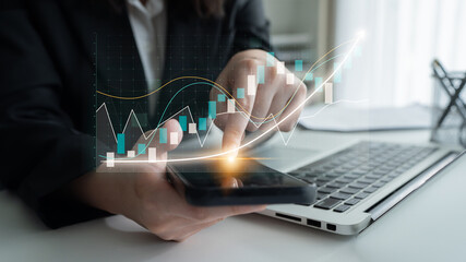 Business professional examines financial data on smartphone with rising stock graphs, using laptop...