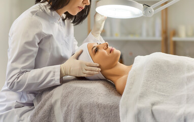 Cosmetologist, dermatologist doing female facial treatment in spa salon, professional magnifying...