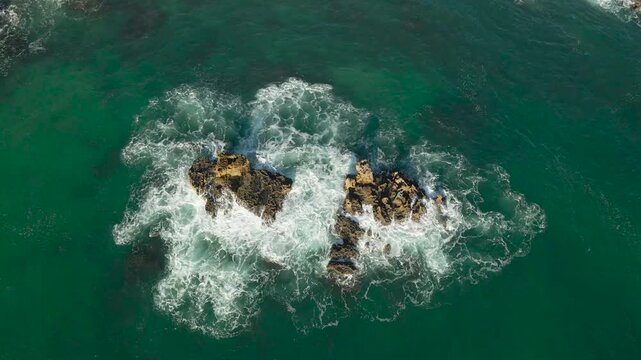 Top down aerial drone video of turquoise green and blue ocean sea water waves hitting a large cliff or a rock karst field in ocean water during a sunny day on Portugal coastline beach rocky shoreline.