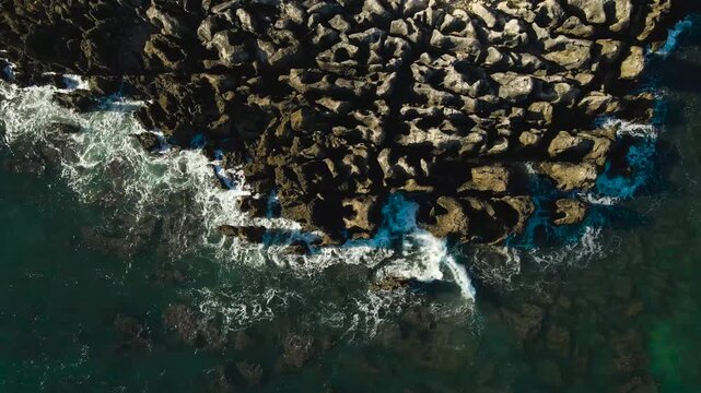 Top down aerial drone video of sharp, tall and dangerous rocky cliffs and rocks on Portugal coastline where foamy white ovean sea water waves hit the rock with high force creating foam and bubbles.