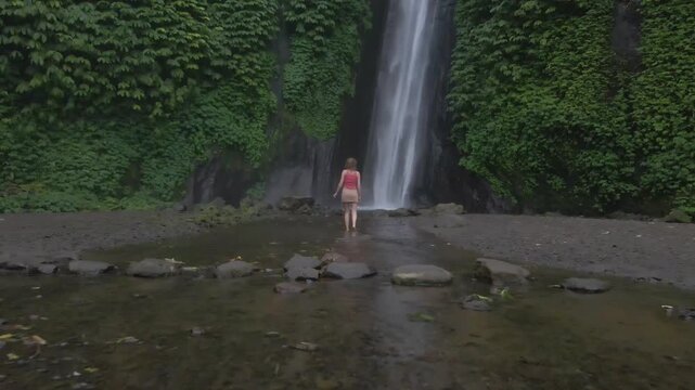 Slow drone video showing woman standing near tall waterfall in lush green forest at Gitgit Falls, Bali, with calm mood and soft mist creating peaceful natural atmosphere.