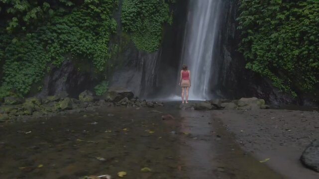 Slow drone video showing woman standing near tall waterfall in lush green forest at Gitgit Falls, Bali, with calm mood and soft mist creating peaceful natural atmosphere.