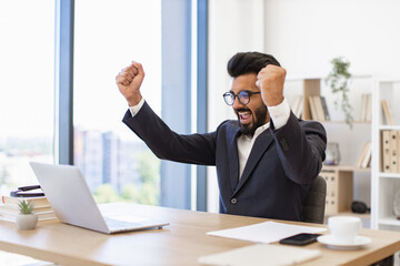 An Indian businessman celebrates a victory in his office, looking at his laptop with excitement