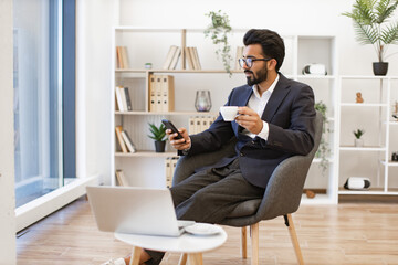 An Indian businessman is relaxing in a modern office setting while checking his phone and enjoying a cup of coffee