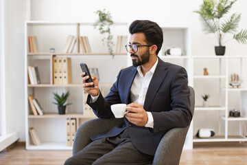 An Arab businessman in a suit is holding a cup of coffee while looking at his phone
