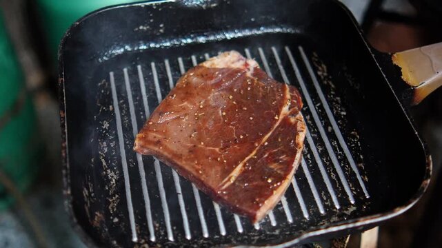 4K raw beef steak is placed on hot grill pan beginning the cooking process with visible searing marks, preparation of grilled meat dish.