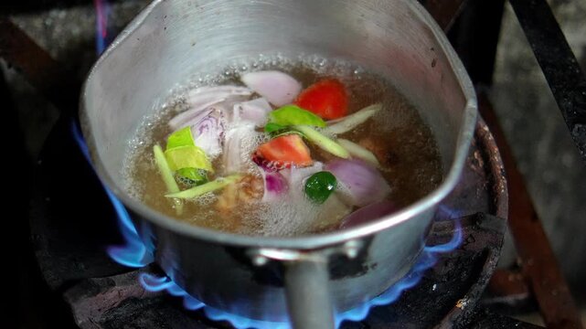 4K pot of traditional Thai herbs including shallots lemongrass kaffir lime leaves galangal and carrots boils over open flame preparation of flavorful soup base.