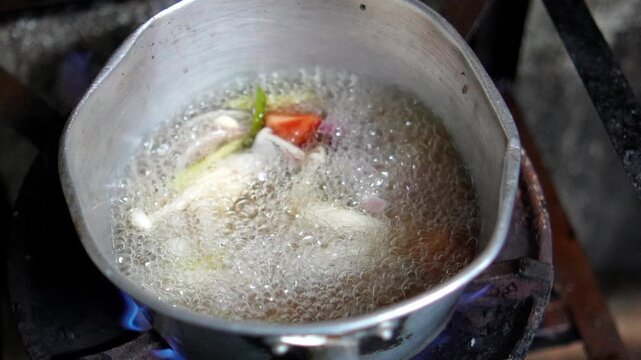 4K pot of traditional Thai herbs including shallots lemongrass kaffir lime leaves galangal and carrots boils over open flame preparation of flavorful soup base.