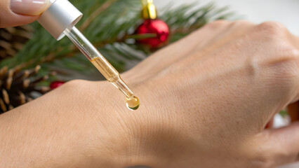 Anti Aging Christmas Skincare A dropper applies serum to the back of a hand with festive pine and ornament decor in the background.