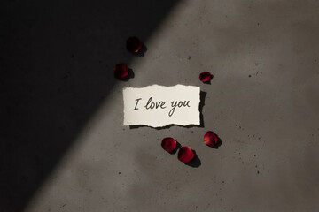 a handwritten i love you note and rose petals on a gray concrete background, top view