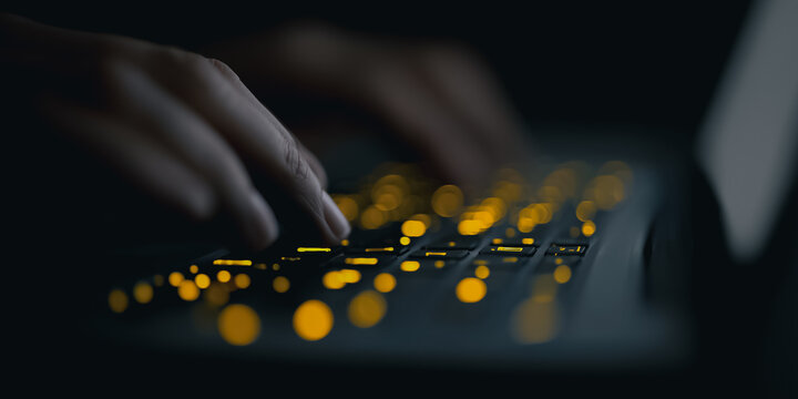 An intimate, low-key view captures fingers poised above a laptop's illuminated keyboard. the warm glow suggests productivity in a dimly lit setting.