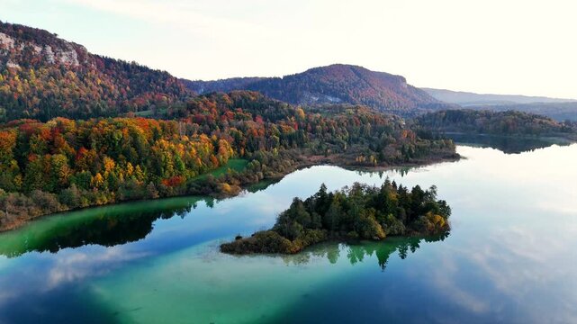 Wide establishing aerial shot of Lac d&rsquo;Ilay in the Jura Mountains, showing the small wooded islet surrounded by turquoise shallows and the autumn‑coloured shoreline