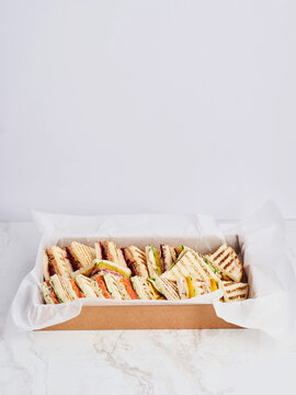 Variety of grilled sandwiches cut into triangles are presented in a brown cardboard box with white parchment paper lining. The sandwiches are on a white marble surface