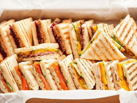 A close-up shows a box filled with a variety of grilled sandwiches. These triangle sandwiches have fillings such as salmon, assorted meats, and cheeses. They are ready to eat
