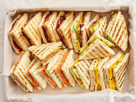 A variety of grilled sandwiches cut into triangles are displayed in a parchment lined tray