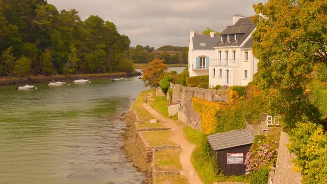 A serene view of the river bank near Le Bono, France, showing traditional white Breton houses, a stone wall covered in autumn ivy, and small boats on the water.