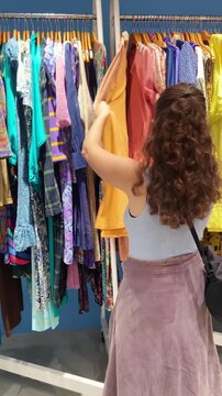 Female looking for second hand clothes and stylish used items on sale in boutique shop or flea market. Young woman browsing clothes on hanger in charity donation retail store. Zero waste influencer