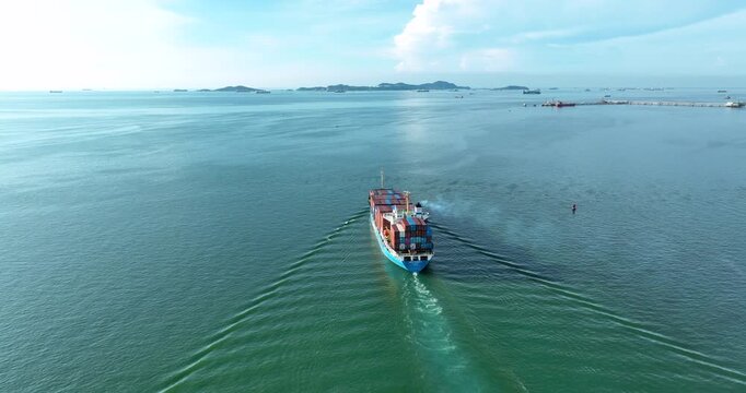 An aerial drone follows a large container ship loaded with cargo as it sails across the open sea, leaving a wake on the water as it heads towards an international commercial shipping port.
