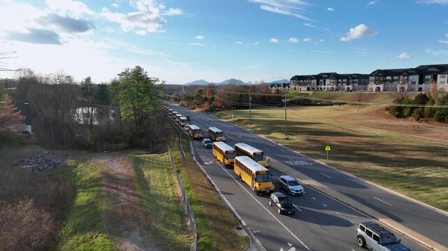 Aerial view of long row yellow school bus moving through America suburb under fall sun, lined with trees, pond reflection and modern apartment complex. blue mountain range in distance.